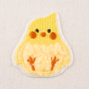 Może przedstawiać: Żółta haftowana łatka przedstawiająca puszystego kurczaka z pomarańczowymi łapkami. Kurczak ma okrągłe, żółte ciało z małym, spiczastym dziobem i czarnymi oczami.
