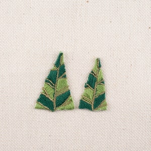 Pode incluir: Dois patches de folhas bordadas em tons de verde e dourado. As folhas têm um design detalhado e texturizado com detalhes em fio dourado. São colocadas sobre um fundo de tecido neutro, branco sujo.