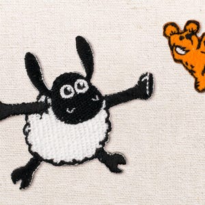 May include: An embroidered patch of a black and white sheep character with outstretched arms and a smiling face, positioned next to a smaller, orange embroidered patch of a dancing bear figure with black outlines and a wide, toothy grin.