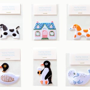 Puede incluir: Seis parches bordados en fundas de plástico transparente individuales con tarjetas de encabezado blancas etiquetadas como HATTA YOSHIE ILLUSTRATION. Los diseños incluyen un perro blanco y negro, una casa azul y rosa, un perro naranja y blanco, un pato, un pingüino y una foca.