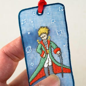 以下が含まれることがあります： 「星の王子さま」のキャラクターが描かれた刺繍のブックマーク。青い背景に白い星が散りばめられています。キャラクターは緑色のコートに赤いアクセントを施し、杖を持っています。上部には赤いリボンが付いています。