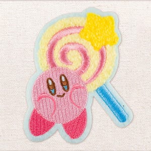 May include: Embroidered patch showing a pink character with a smiling face and large eyes, holding a yellow, pink, and blue lollipop. The patch is outlined in light blue and is on a white fabric background.