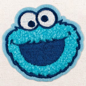 Op de afbeelding: Geborduurde patch met het gezicht van Koekiemonster. Het ontwerp omvat grote witte ogen met zwarte pupillen, een wijd open mond en een blauw, getextureerd lichaam. De patch is omrand in een donkerdere blauwe kleur.