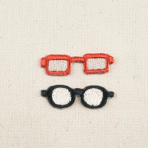 以下が含まれることがあります： ベージュの布地に刺繍された眼鏡が2組。上の組は赤でレンズが白。下の組は黒でレンズが白です。