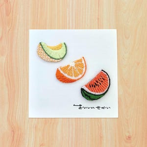 May include: Embroidered fruit slices on a white square. The image features a cantaloupe, orange, and watermelon slice. The cantaloupe is light green and yellow, the orange is orange and white, and the watermelon is red and green. Japanese text is at the bottom.
