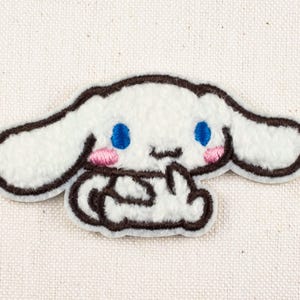 Peut inclure: Un écusson en peluche blanche représentant un personnage de dessin animé avec de grandes oreilles, des yeux bleus et des joues roses. Le personnage a un contour marron et est posé sur un fond en tissu neutre. Idéal pour les vêtements ou accessoires.