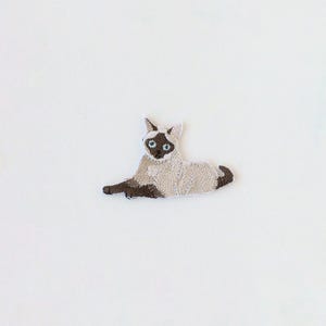 May include: An embroidered iron-on patch featuring a Siamese cat in a reclining position. The cat has a cream-coloured body, dark brown ears, face, and paws, and bright blue eyes. The embroidery details show the cat's fur texture and features.