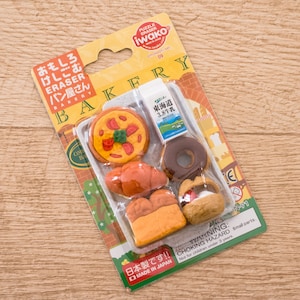 May include: A pack of six small eraser toppers shaped like bakery items, including a pizza, a croissant, a loaf of bread, a doughnut, and a milk carton. The packaging is yellow and brown with Japanese text that reads "Omoshiro Keshigoma Pan Yasan" which translates to "Fun Erasers Bakery".