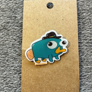 May include: A teal blue cartoon platypus wearing a black cowboy hat with a rainbow band. The platypus has large, white eyes and a yellow beak. The platypus is standing on a white background with the text "FUNZIGG" printed below it.