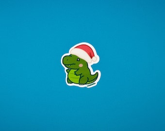 May include: A green dinosaur sticker wearing a red and white Santa hat. The sticker has a white border and is set against a bright blue background. The dinosaur has a friendly expression.