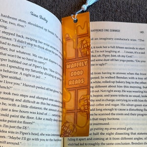May include: A bookmark with a waffle design and the text "Waffely Good Reads" in orange and white. The bookmark is made of a clear plastic material and has a tassel.