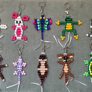 Puede incluir: Una colección de diez llaveros con forma de animal hechos de cuentas de plástico de colores. Las figuras incluyen una abeja, un conejo rosa, una mariposa, una rana verde, un gato, un mono, un lagarto, una nutria, una criatura marrón y una tortuga verde.