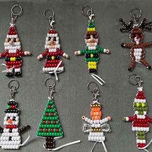 May include: A collection of eight festive-themed keychains crafted from colourful plastic pony beads. The figures include Santa Claus, an elf, a reindeer, a snowman, a Christmas tree, an angel, and a green character, each with a metal keyring.