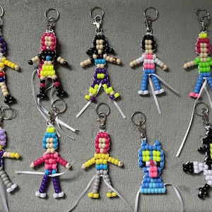 May include: A collection of nine handmade keychains crafted from colorful plastic pony beads. Eight keychains feature human-like figures with various hair and clothing colors, while one keychain depicts a blue cat-like character and another a black and white creature.