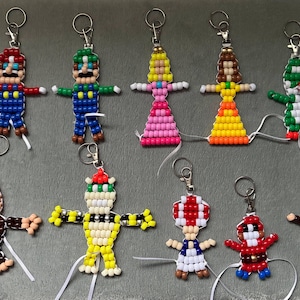 May include: A collection of ten handmade keychains crafted from colourful plastic pony beads, depicting various video game characters including Mario, Luigi, Princess Peach, Daisy, Yoshi, Donkey Kong, Bowser, Toad, a red-clad figure, and Diddy Kong.