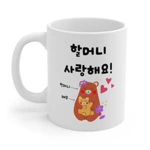 May include: White ceramic mug with a brown bear and a smaller brown bear cub hugging. The text "할머니 사랑해요!" is written in black on the mug. The text "할머니" is written in black with an arrow pointing to the larger bear. The text "DHF듀" is written in black with an arrow pointing to the smaller bear. There are pink hearts and purple flowers on the mug.