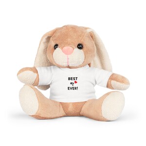 May include: A plush bunny rabbit with tan fur, a pink nose, and black eyes. The bunny wears a white t-shirt with the words "BEST 💖 EVER!" printed in black and red. The bunny is sitting upright.