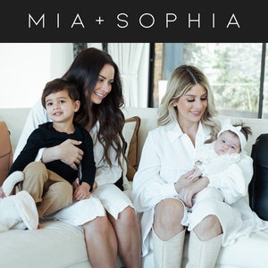 May include: Two women are sitting on a white couch with their children. One woman is holding a baby and the other is holding a toddler. Both women are wearing white shirts and the children are wearing casual clothing. There are two leather diaper bags on the couch, one brown and one black.