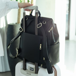 May include: A black leather backpack with adjustable straps and a zippered pocket is attached to a white suitcase with a brown trim. The suitcase has a retractable handle.