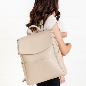 May include: A beige backpack with a flap closure, side pockets, and a top handle. The bag is made of a smooth material and has a minimalist design. The backpack is being carried by a person.