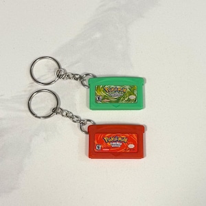 May include: Two keychains featuring miniature Game Boy Advance cartridges. One is green with "Pokemon LeafGreen Version" text, the other is red with "Pokemon FireRed Version" text. Each keychain has a silver chain and ring.