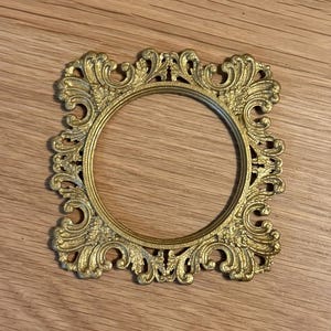 Könnte beinhalten: Verzierter, goldfarbener, kreisförmiger Bilderrahmen mit detailliertem, geschwungenem Design. Der Rahmen ist aus Metall und hat eine Vintage-Ästhetik. Der Rahmen ist leer und bereit, ein Foto oder Kunstwerk aufzunehmen.