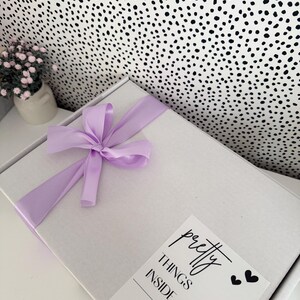 Pu&ograve; includere: Una scatola regalo rettangolare bianca legata con un fiocco di nastro di raso viola chiaro. Un'etichetta bianca sulla scatola presenta il testo nero "pretty THINGS INSIDE" e "OPEN ME" con piccole icone a forma di cuore nero.