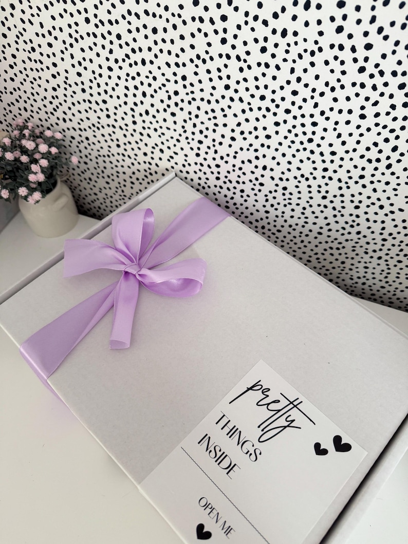 Pu&ograve; includere: Una scatola regalo rettangolare bianca legata con un fiocco di nastro di raso viola chiaro. Un'etichetta bianca sulla scatola presenta il testo nero "pretty THINGS INSIDE" e "OPEN ME" con piccole icone a forma di cuore nero.