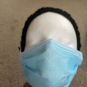 May include: A light blue disposable face mask is on a white mannequin head. The mask is pleated with a white border. A black hair band is around the mannequin head.