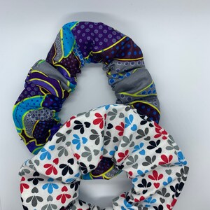 May include: Two fabric scrunchies. One is patterned with purple, blue, and yellow geometric shapes. The other is white with a pattern of red, blue, and black leaf shapes. Both are circular and appear to be made of cotton.
