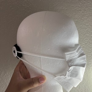 May include: White face mask with ear loops, attached to a black fabric piece with a white button. The mask is on a white mannequin head. The mask is pleated and has a rectangular shape.