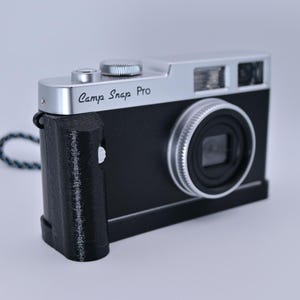 May include: A black and silver camera with the text "Camp Snap Pro" on the top. The camera has a textured grip and a lens with a silver ring. A blue and gray strap is attached to the side.