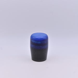 May include: A small, cylindrical object with a smooth, rounded top. The surface features a gradient color transition, starting with a vibrant blue at the top, blending into a deep royal blue in the middle, and finishing with a dark black base.