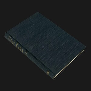 May include: A hardcover book with a dark teal cover and gold lettering on the spine. The book is closed and angled, with a textured appearance. The title and author's name are visible on the spine. The pages are white.
