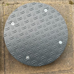 Può includere: Un coperchio di accesso rotondo in metallo nero con un motivo a piastra diamantata. Il coperchio è fissato con quattro viti.