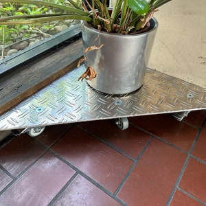 Può includere: Un portavasi triangolare in argento con ruote, con un motivo a rombi. Il supporto sostiene un vaso in argento con una pianta verde.