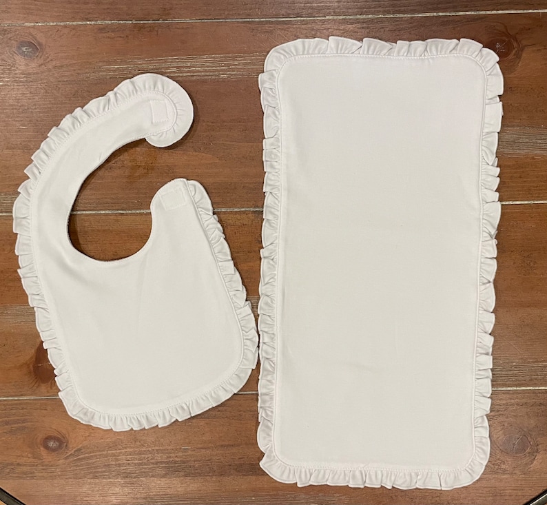 May include: A white baby bib and changing mat set. Both items are rectangular and have a ruffled edge. The bib has a curved neck opening and a Velcro fastening. The changing mat is a simple rectangle. The set is made of a soft, white fabric.
