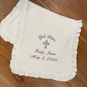 May include: A white baby blanket with a frilled border. The blanket has embroidered text in purple thread: "God Bless", a cross, the name "Ruby Jean", and the date "May 3, 2026". The blanket is folded on a wooden surface.
