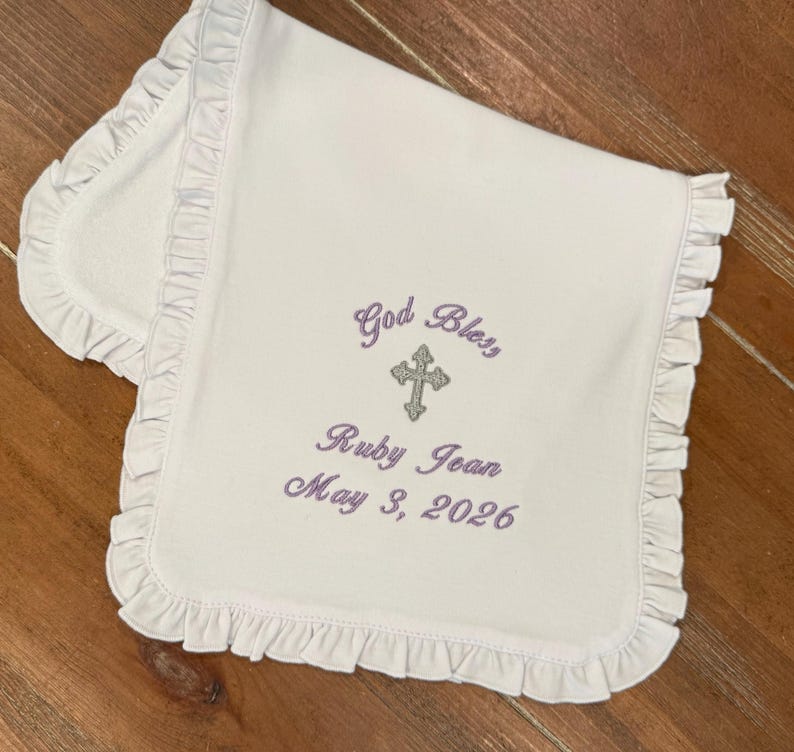 May include: A white baby blanket with a frilled border. The blanket has embroidered text in purple thread: "God Bless", a cross, the name "Ruby Jean", and the date "May 3, 2026". The blanket is folded on a wooden surface.