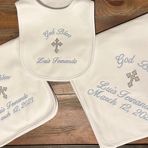 May include: A set of white baby items, including a bib and two blankets, embroidered with the words "God Bless", a cross, and the name "Luis Fernando". One blanket is dated "March 12, 2023". The items are displayed on a wooden surface.