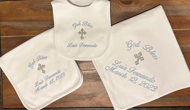 May include: A set of white baby items, including a bib and two blankets, embroidered with the words "God Bless", a cross, and the name "Luis Fernando". One blanket is dated "March 12, 2023". The items are displayed on a wooden surface.