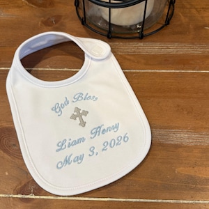 May include: White baby bib with blue embroidered text that reads "God Bless, Liam Henry May 3, 2026" and a cross. The bib has a rounded shape and a snap closure at the neck.