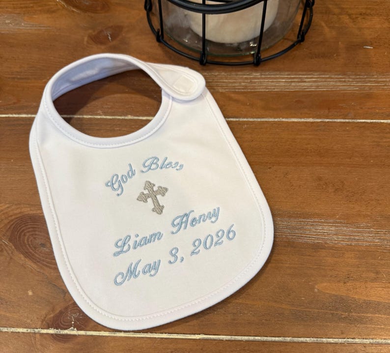 May include: White baby bib with blue embroidered text that reads "God Bless, Liam Henry May 3, 2026" and a cross. The bib has a rounded shape and a snap closure at the neck.