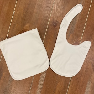 May include: Two white baby bibs on a wooden surface. One is rectangular, and the other has a curved shape with a snap closure. Both are made of a soft, smooth material and have a simple, clean design.