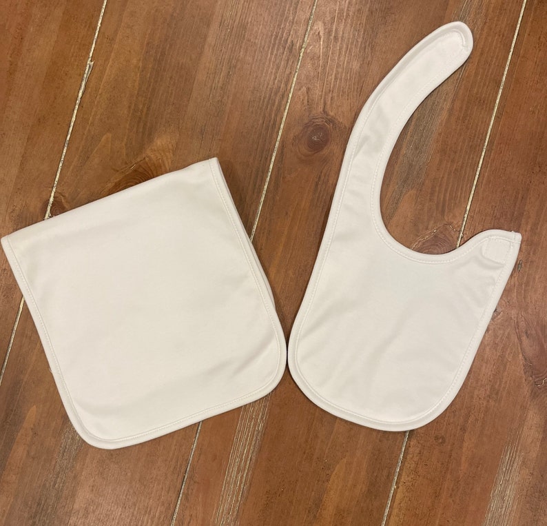 May include: Two white baby bibs on a wooden surface. One is rectangular, and the other has a curved shape with a snap closure. Both are made of a soft, smooth material and have a simple, clean design.