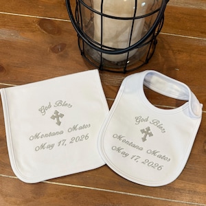May include: A white baby bib and matching burp cloth. Both are embroidered with the words "God Bless", a cross, the name "Montana Matos", and the date "May 17, 2026" in grey thread. The bib has a snap fastening.