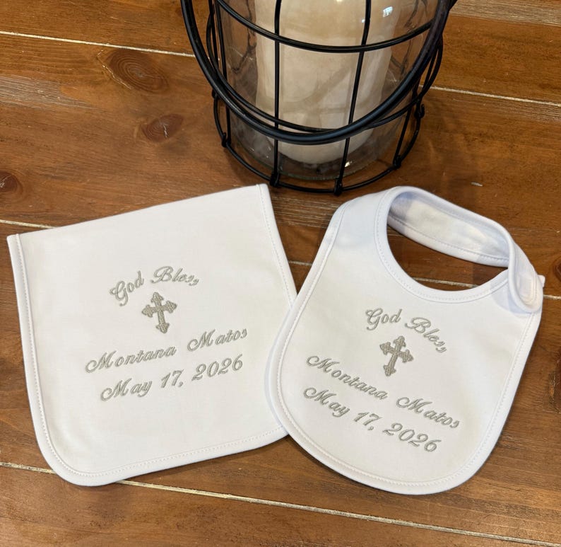 May include: A white baby bib and matching burp cloth. Both are embroidered with the words "God Bless", a cross, the name "Montana Matos", and the date "May 17, 2026" in grey thread. The bib has a snap fastening.