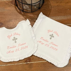 May include: White bib and burp cloth set with frilled edges. Both items are embroidered with "God Bless", a cross, the name "Emilia James", and the date "May 23, 2026" in pink thread.