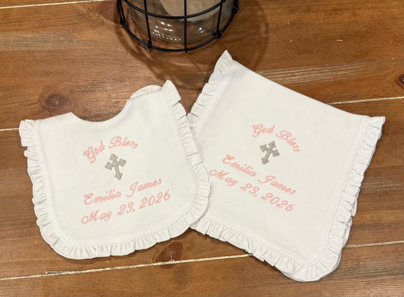 May include: White bib and burp cloth set with frilled edges. Both items are embroidered with "God Bless", a cross, the name "Emilia James", and the date "May 23, 2026" in pink thread.