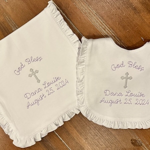 May include: White baby burp cloth and bib with ruffled edges. Both items are embroidered with the text "God Bless", a cross, "Dana Louise", and "August 25, 2024".